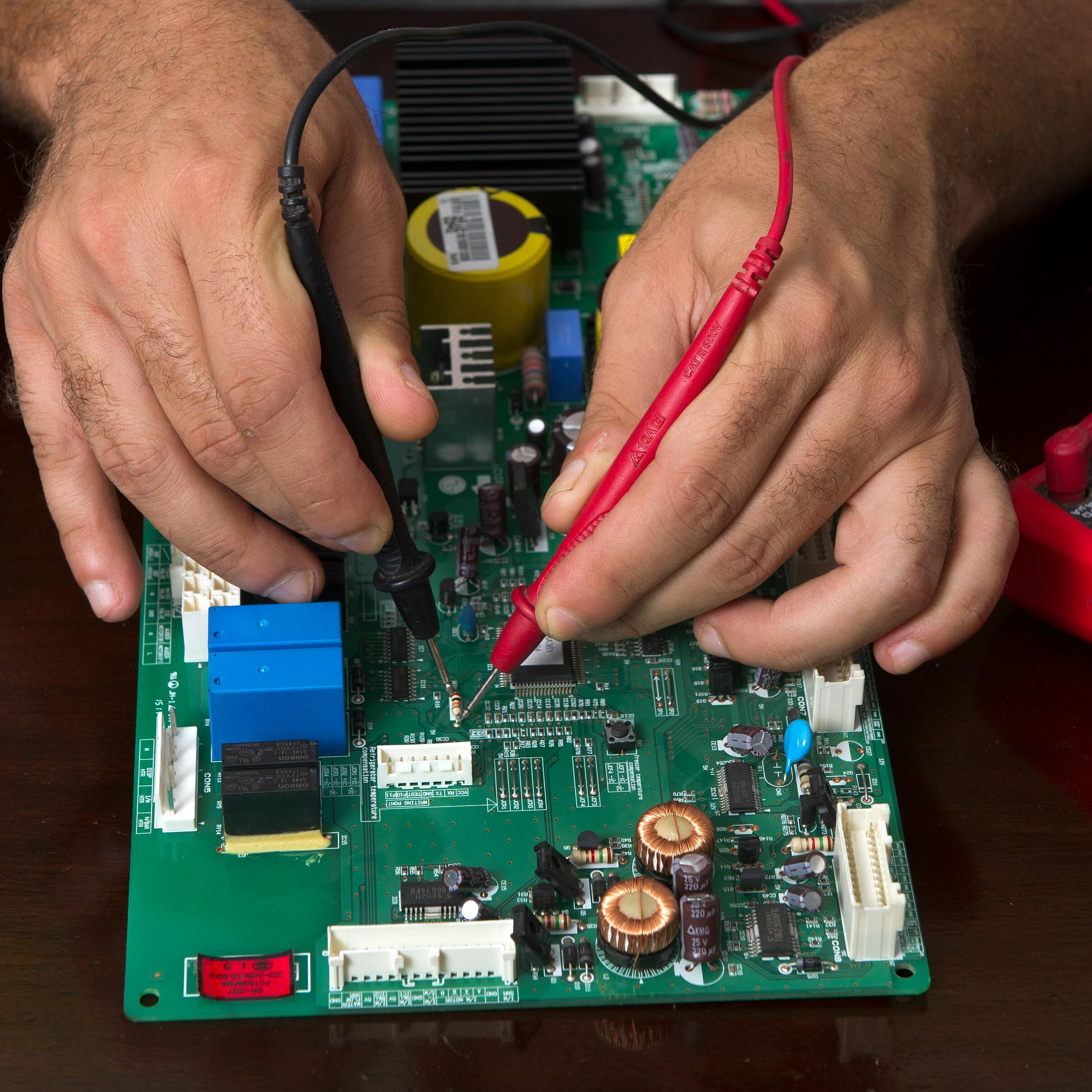 Hands using red and black probes to test a green circuit board with various electronic components on a dark surface.