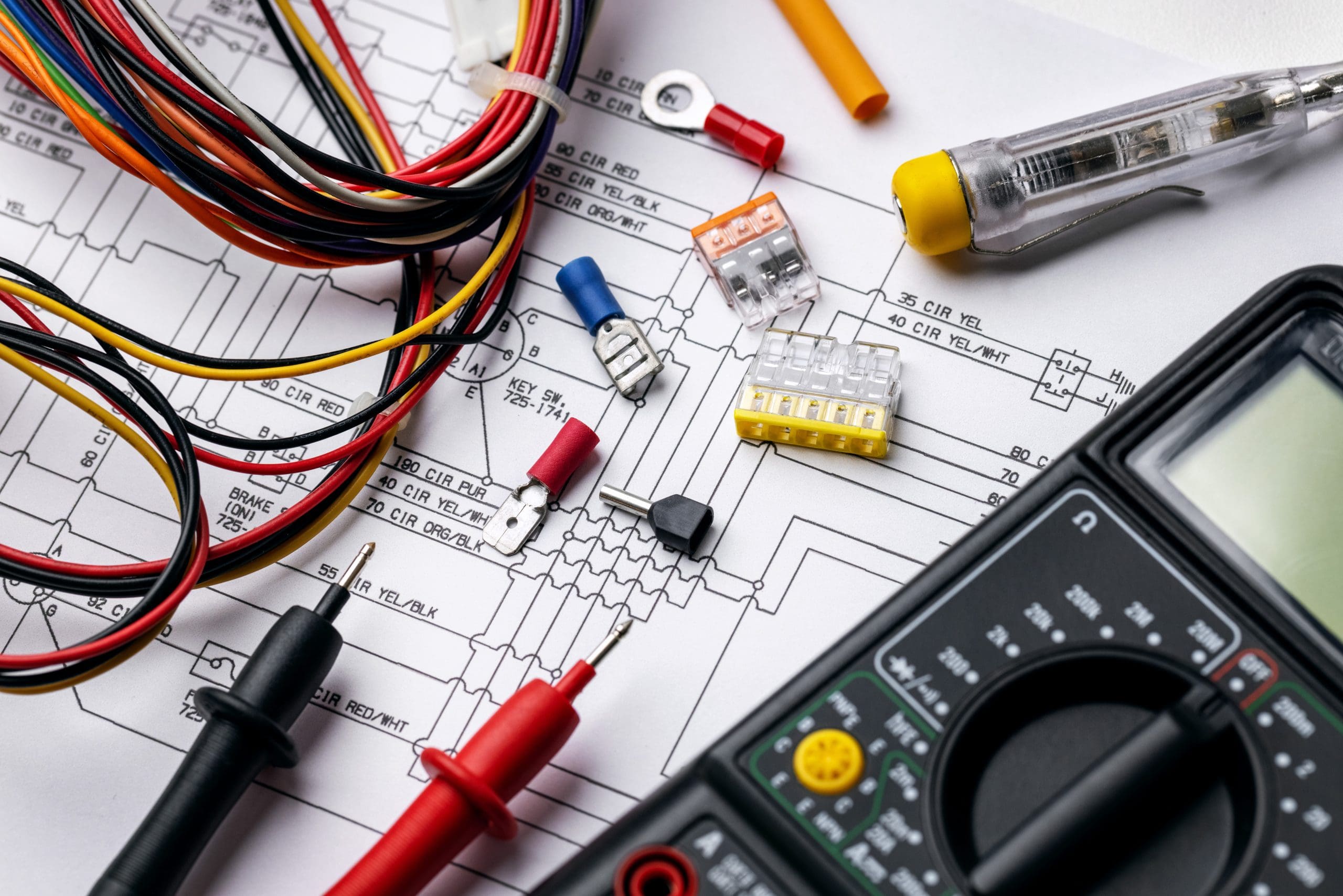 A multimeter and test leads rest on an electrical circuit diagram, surrounded by various electronic components such as wires, fuses, and connectors.