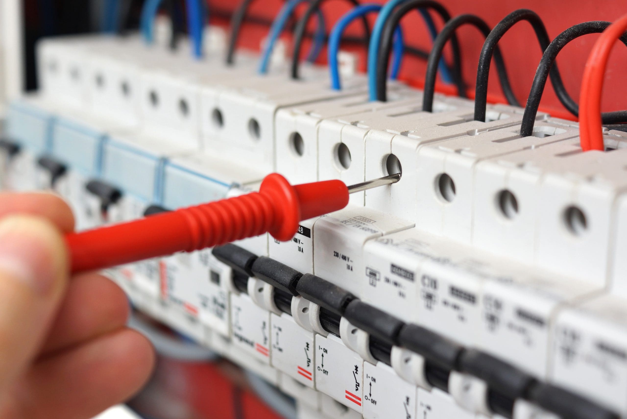 A hand holding a red insulated screwdriver is adjusting electrical connections in a circuit breaker panel with multiple wires and switches, showcasing electrical maintenance or installation work.