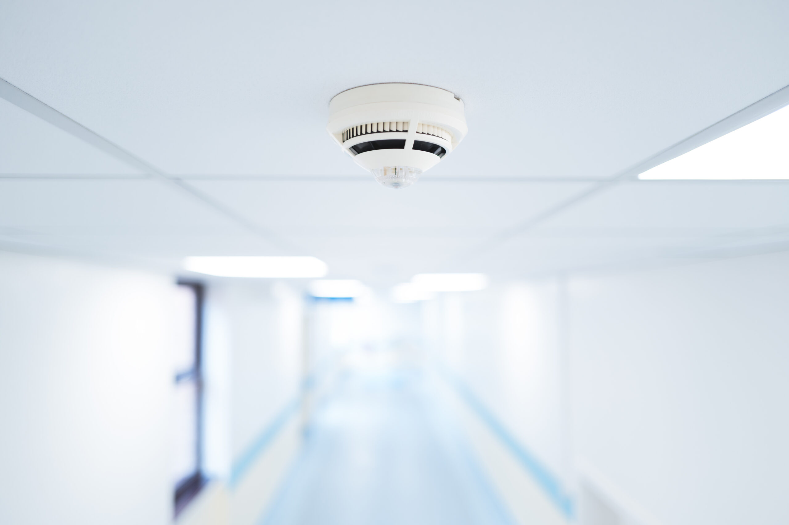 A close-up of a white smoke detector mounted on the ceiling of a bright, empty hallway with white walls and blue accents. The background is blurred, emphasizing the safety device.