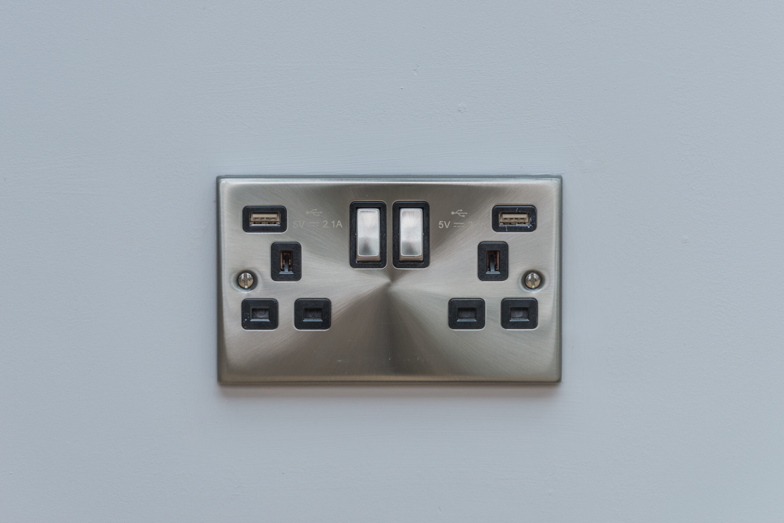 A brushed metal double electrical wall socket with two on/off switches and two USB ports, mounted on a plain light gray wall.