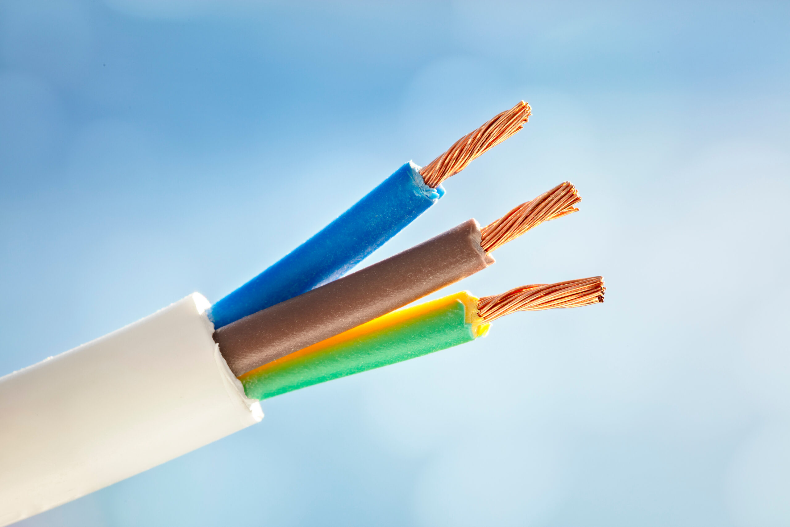 Close-up of three exposed copper wires inside a white electrical cable; the wires are individually covered with blue, brown, and green-yellow insulation, set against a blurred blue background.