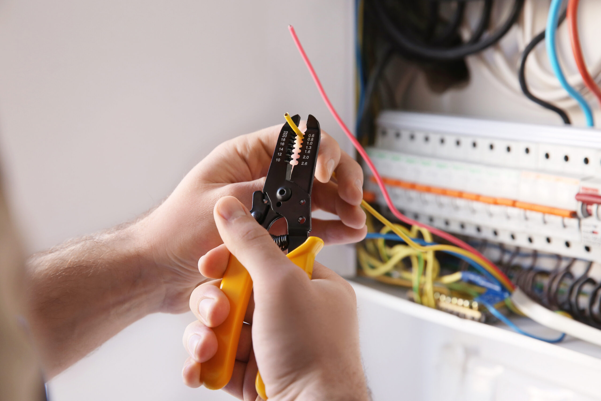 A person uses a yellow-handled wire stripper to strip insulation from a red electrical wire near an open electrical panel with various colored wires inside.