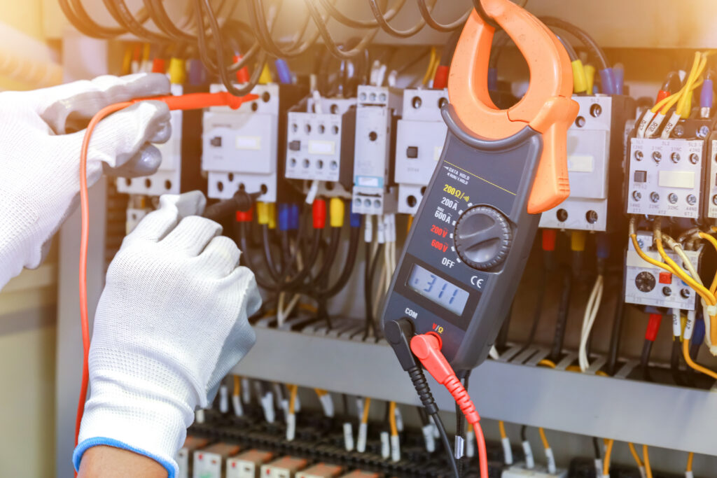 A person wearing white gloves uses a digital clamp meter to measure voltage or current in an industrial electrical control panel with multiple wires and circuit breakers.