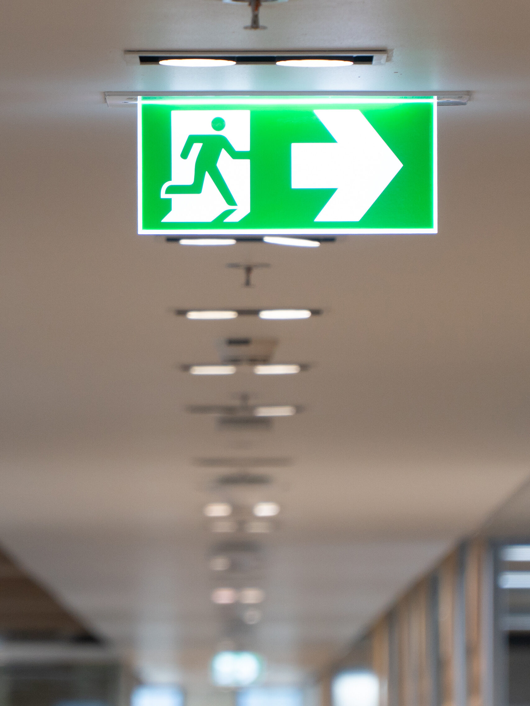 A green emergency exit sign with a running figure and a large white arrow pointing right, mounted on a ceiling in a hallway with modern lighting.