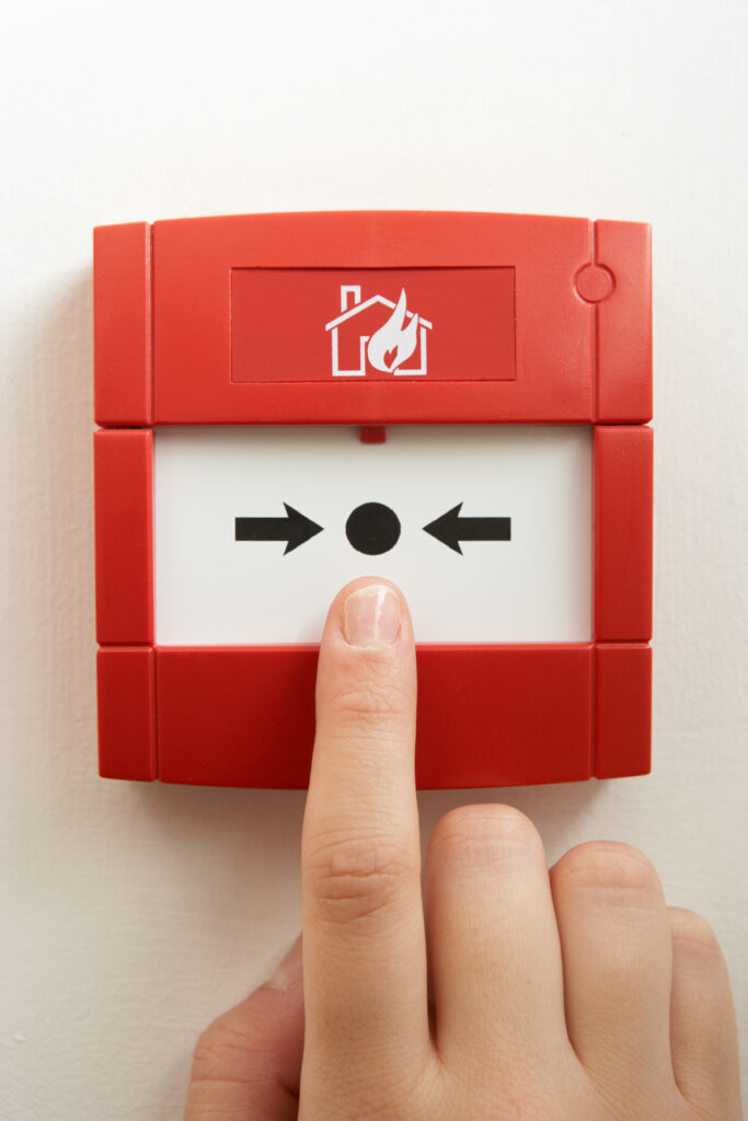 A hand pressing a red fire alarm button mounted on a white wall, with black arrows pointing toward a central black dot and a fire symbol above.